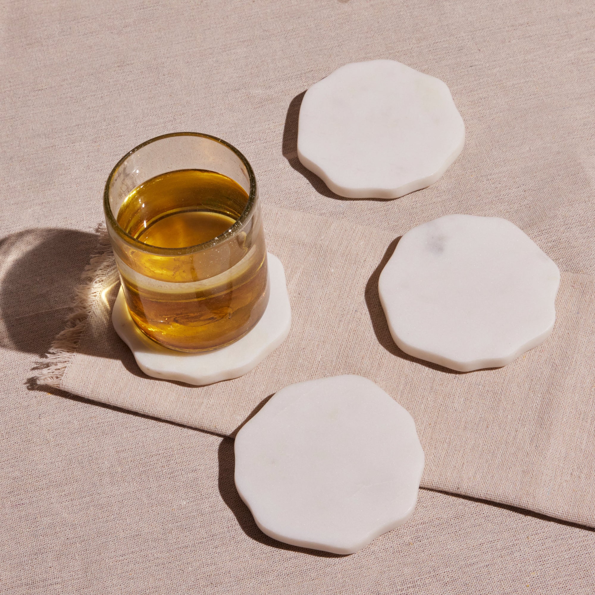 A set of four white, octagonal marble coasters with a glass of amber liquid on one of them, displayed on a beige surface
