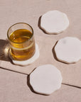 A set of four white, octagonal marble coasters with a glass of amber liquid on one of them, displayed on a beige surface