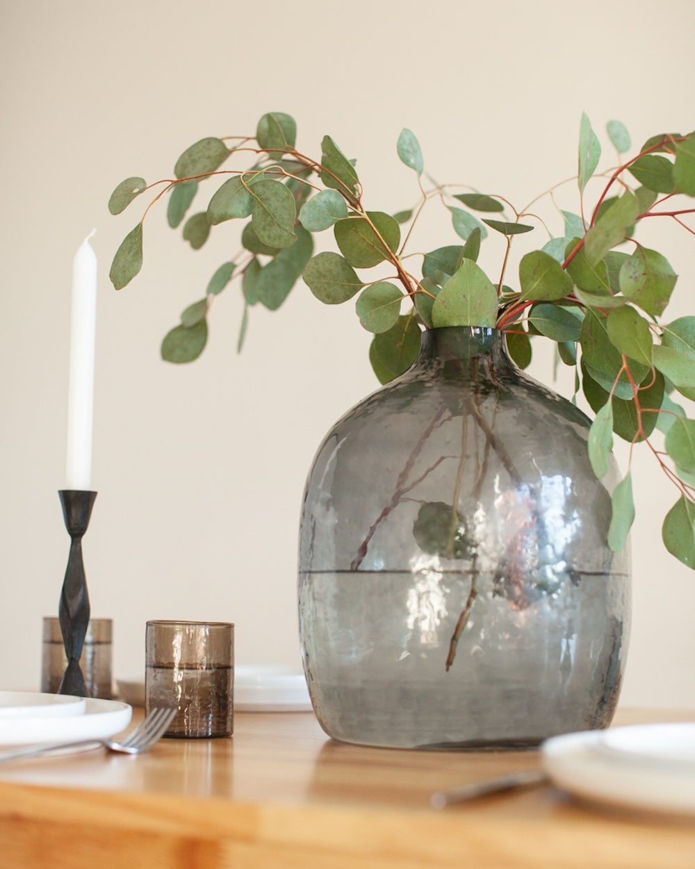 Decorative setting with a vase of greenery, candle, and cutlery on a table.