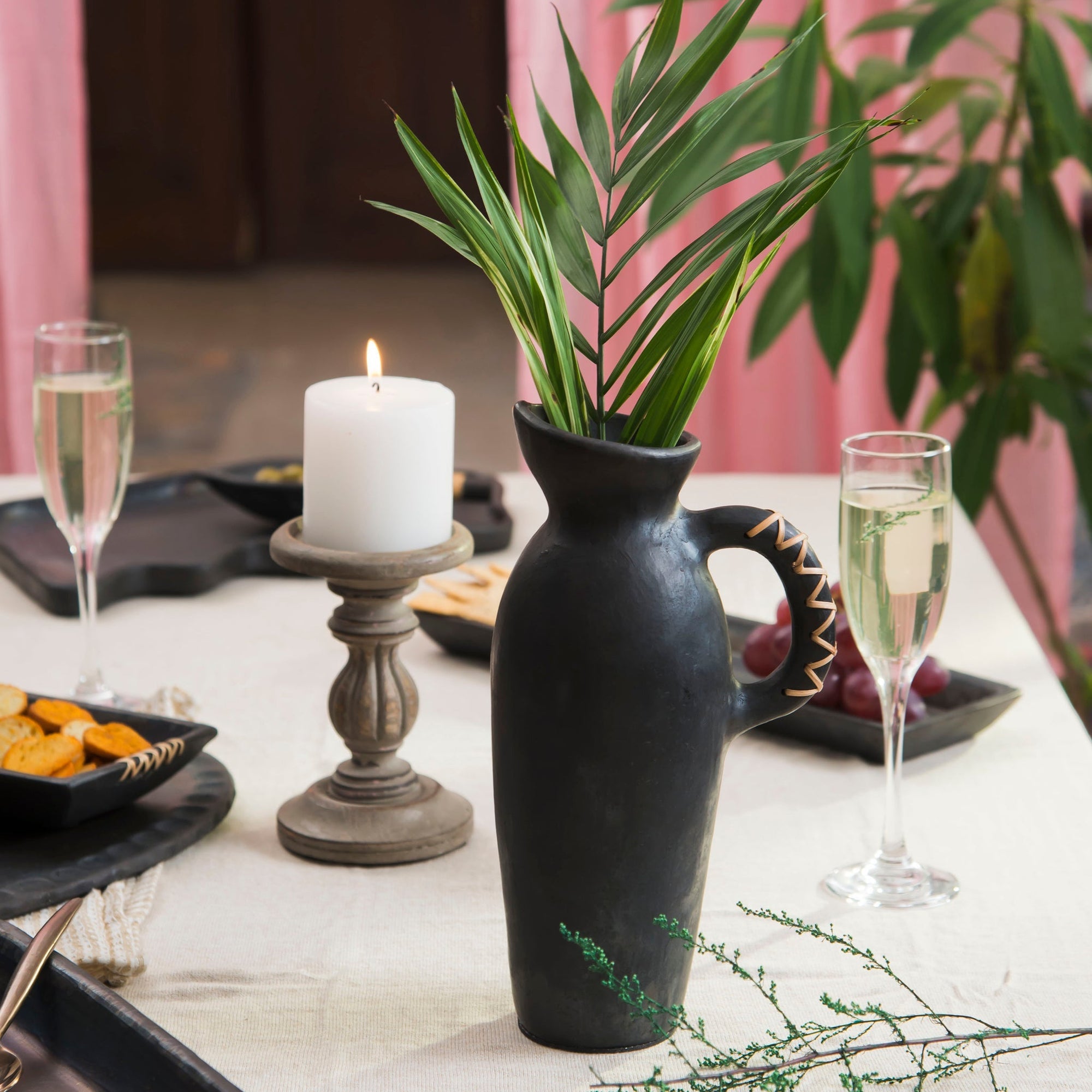 A black ceramic vase with a gold geometric pattern on the handle with plants inside on a table near wine glasses and a candle