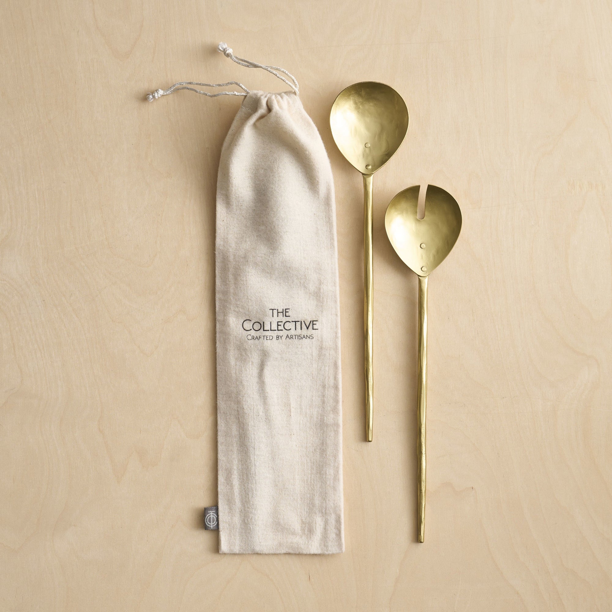 A set of two brass salad servers, a fork and a spoon, with long handles, displayed next to a beige flannel storage bag with the brand 'The Collective' visible on it