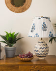 Decorative lamp with floral shade on a wooden table with books, grapes, and a plant.