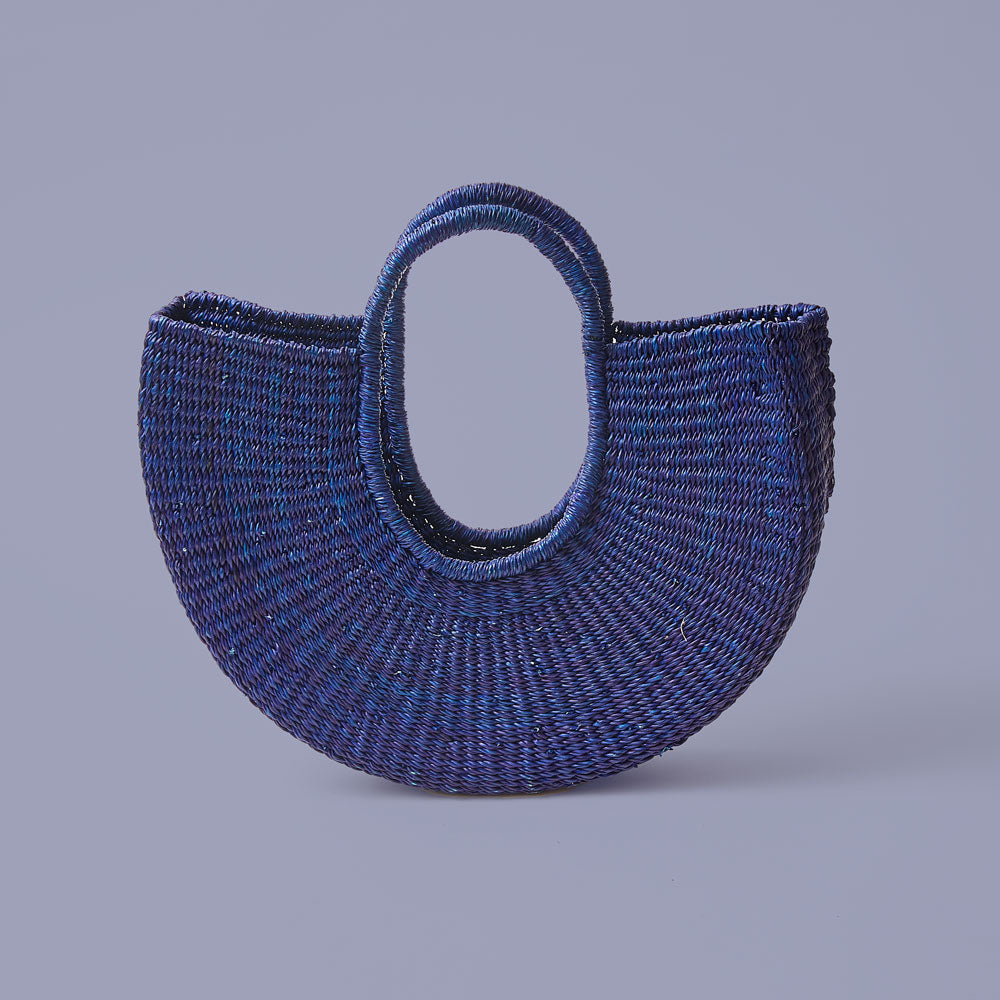 A hand-dyed blue, half-moon shaped handbag made from elephant grass, with an open top and two handles.