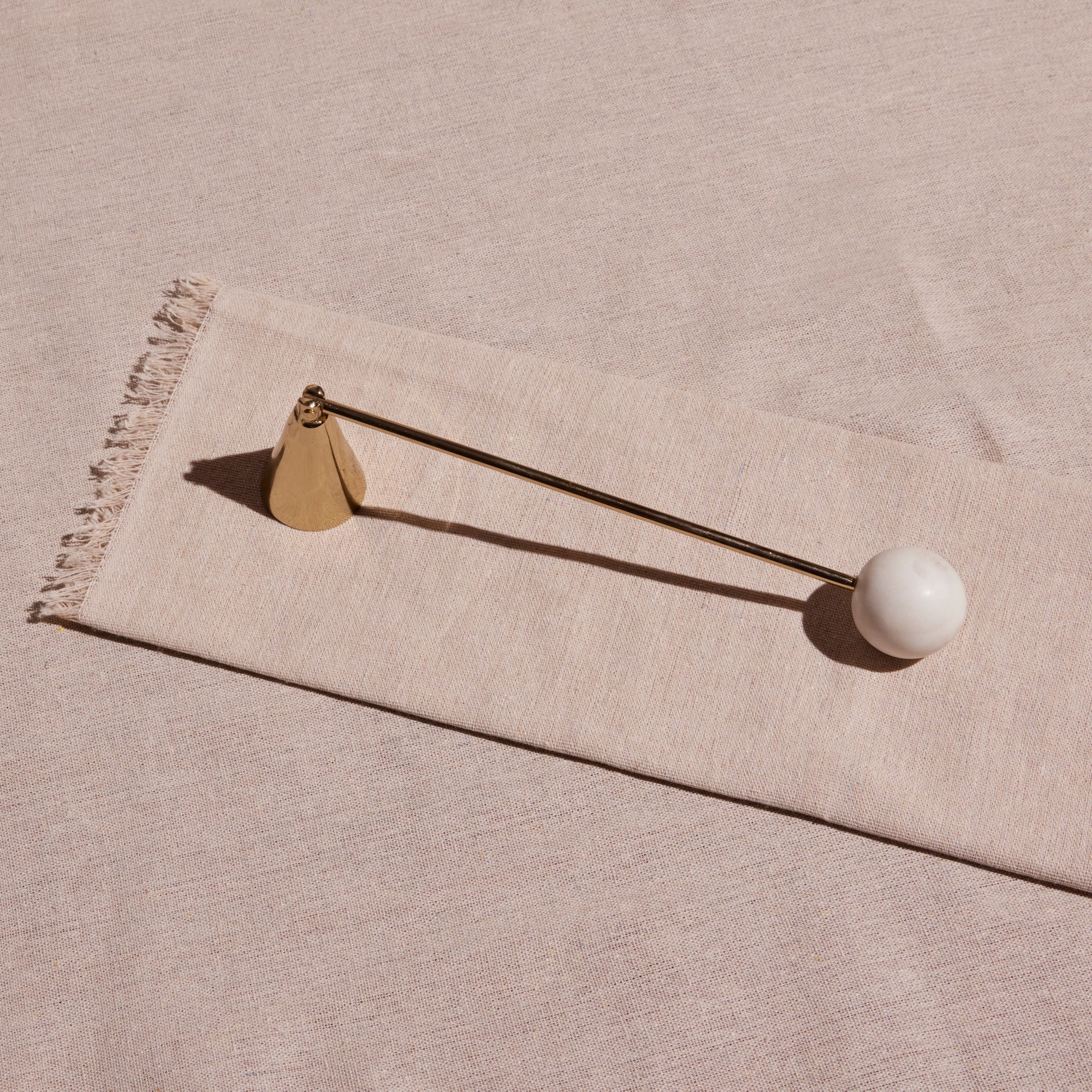 A stainless steel candle snuffer with a brass finish and white marble detail, laid on a beige textile surface.
