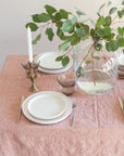 Table setting with white plates, silverware, and a vase with greenery on a pink tablecloth.