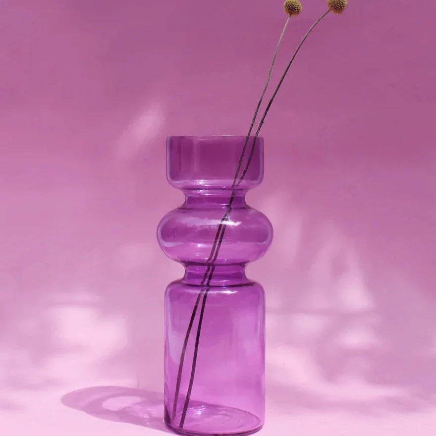 A purple glass vase with a translucent finish, featuring a symmetrical design, filled with dry twigs, positioned against a neutral background
