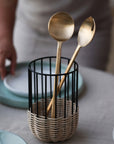 A pair of brass salad server in a black wire holder on a table with a blurred background