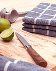 Two terry kitchen towels on a wooden cutting board next to a knife and limes