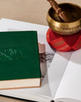Green book with a lotus embossment next to a bronze singing bowl on a red cushion.