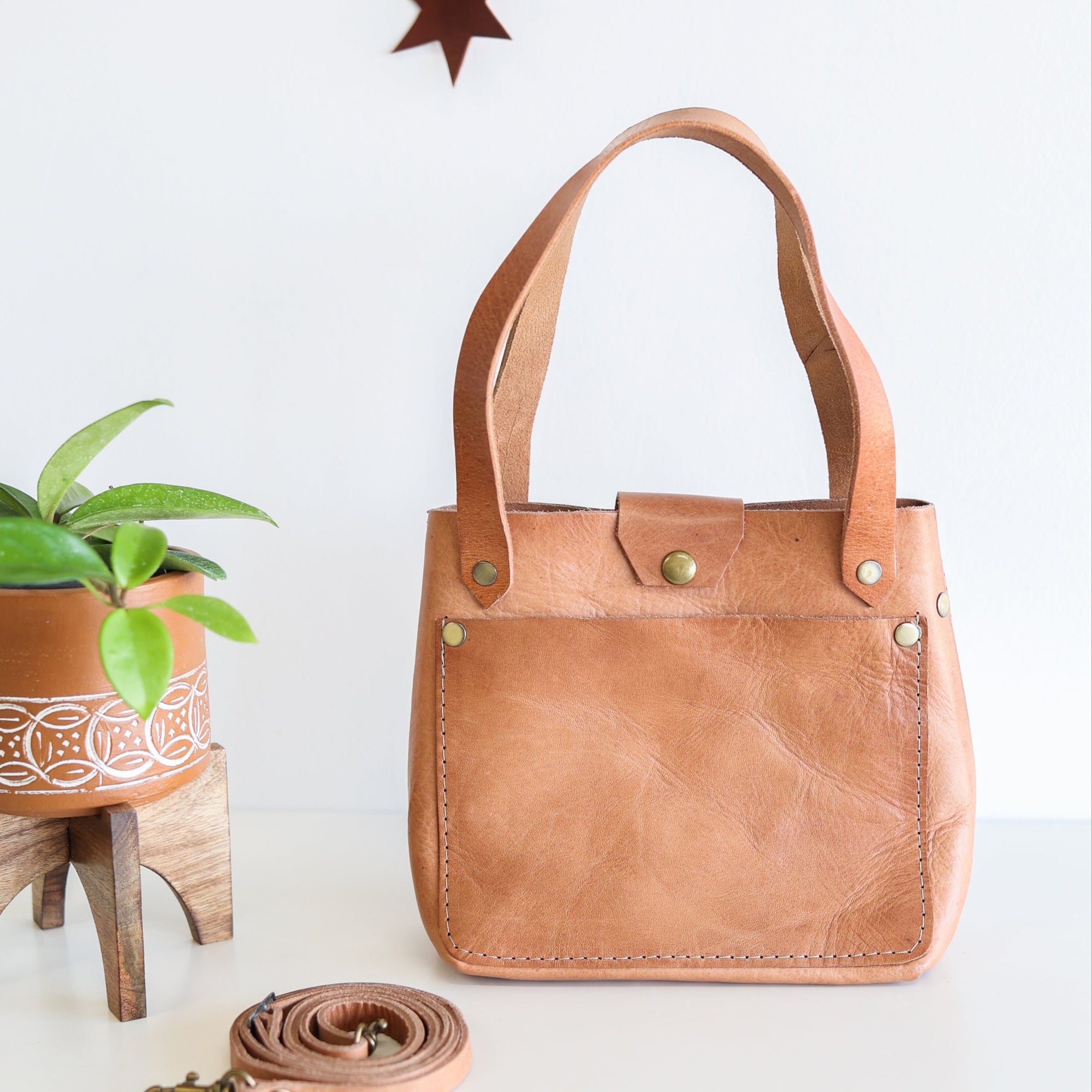 A brown full grain leather mini crossbody tote with a handle and a front pocket, displayed next to a plant pot.
