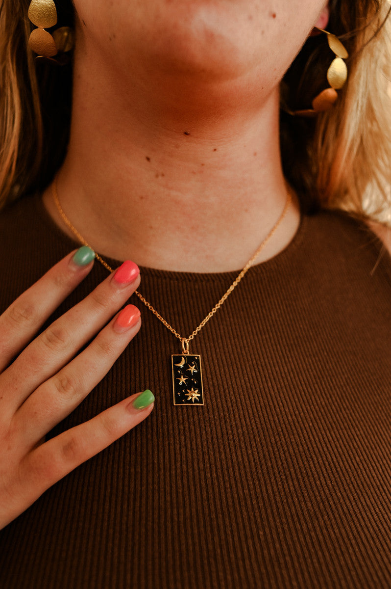 A model wearing a gold-plated pendant necklace with a rectangular black enamel pendant featuring a white star and moon pattern, connected to a chain by a small gold-plated bar.