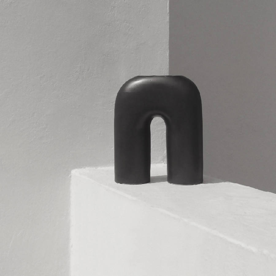 A black, sculptural, inverted-U-shaped vase placed on a white surface in a minimalistic setting.