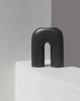 A black, sculptural, inverted-U-shaped vase placed on a white surface in a minimalistic setting.