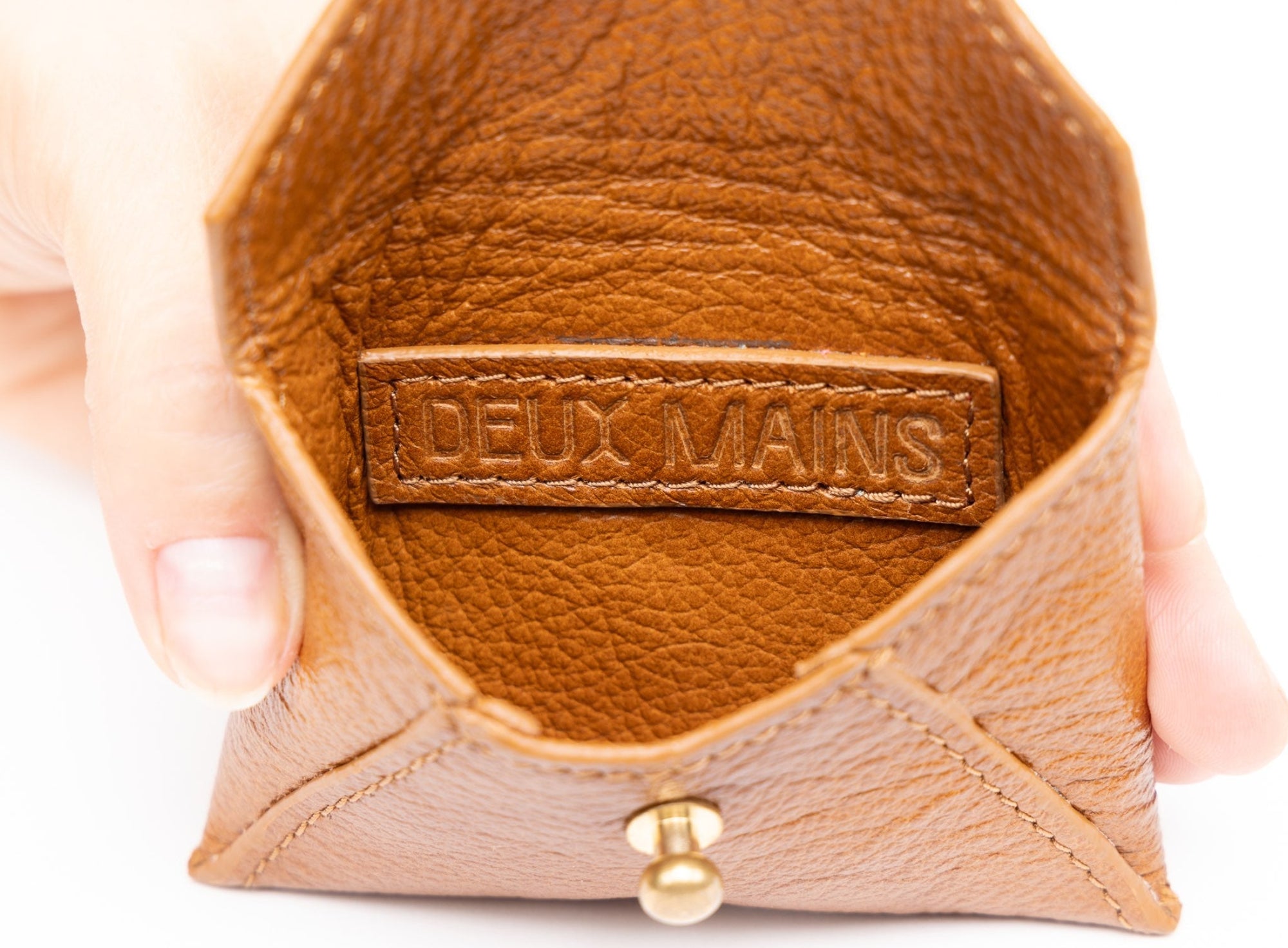 Brown leather product with 'DEUX MAINS' label held by a hand on a white background