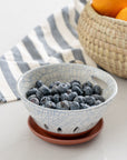 Ceramic bowl with blueberries on a white surface with a striped cloth and basket of oranges in the background.