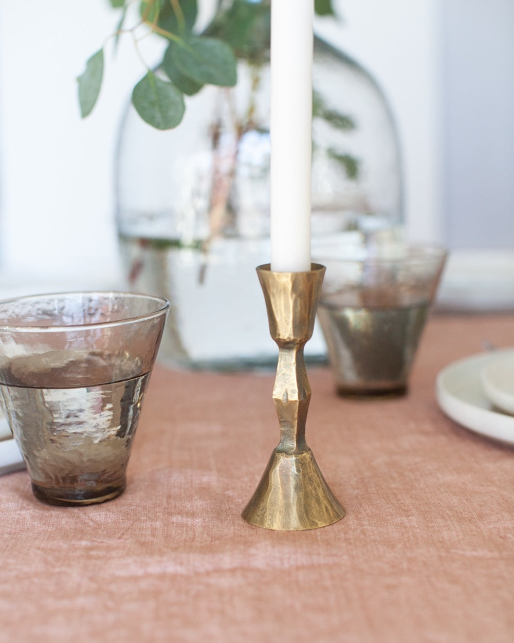 Gold candle holder on a table with glasses and a plant in the background