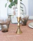 Gold candle holder on a table with glasses and a plant in the background