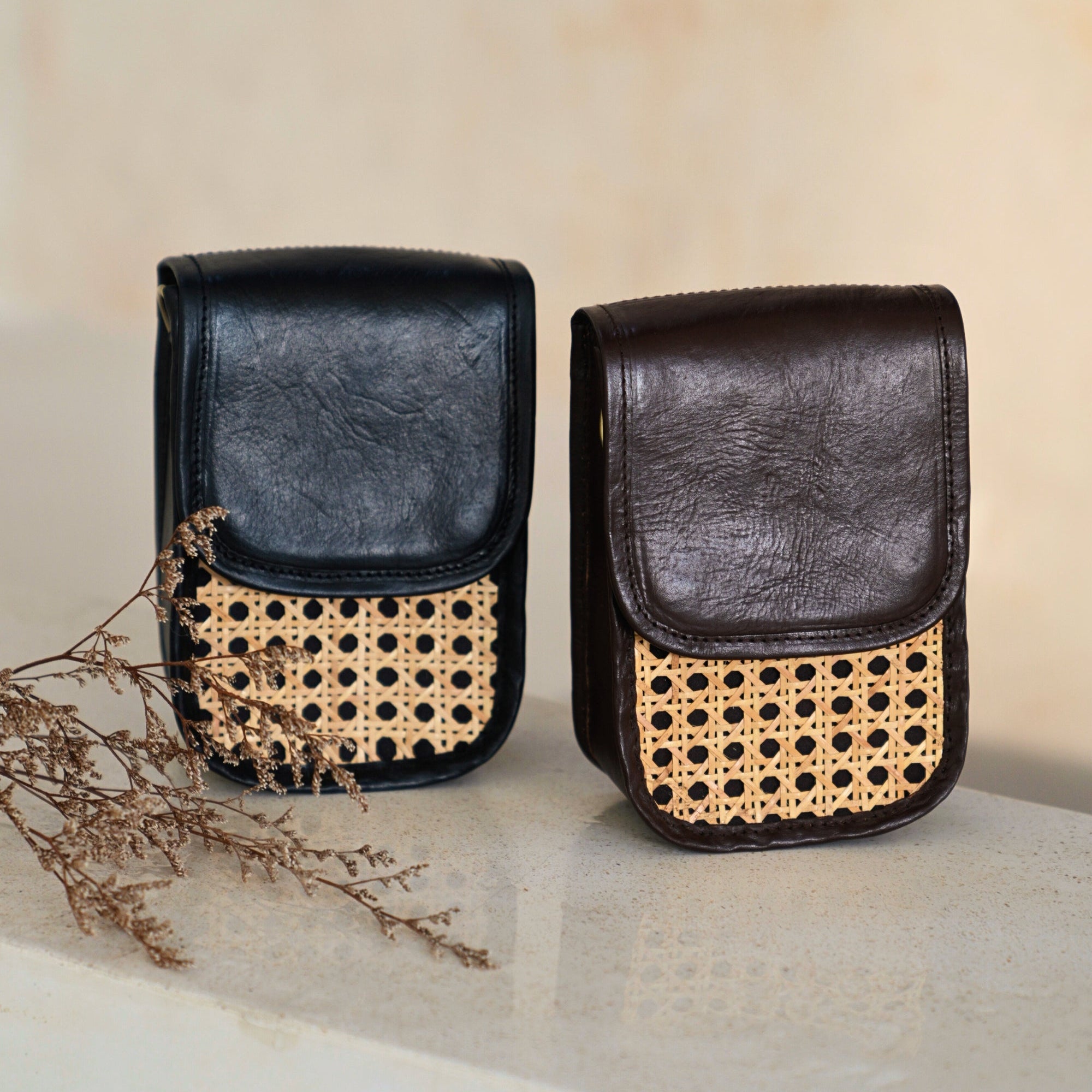 One black and one brown clutch crossbody bags with rattan cane pattern on the front and leather trim, featuring a detachable shoulder strap on a marble table.