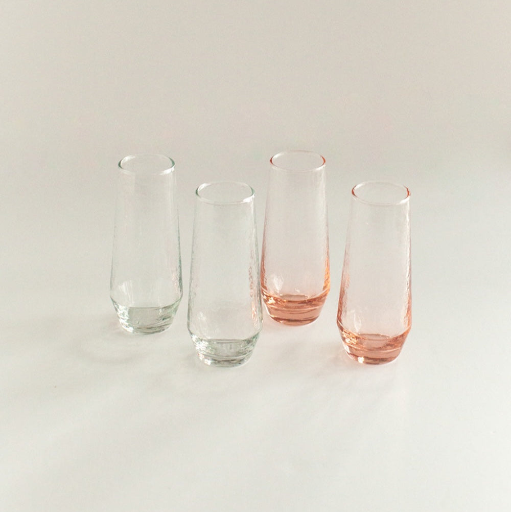 A set of four stemless champagne flutes, three clear and one with a pink hue, displayed against a neutral background