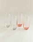 A set of four stemless champagne flutes, three clear and one with a pink hue, displayed against a neutral background