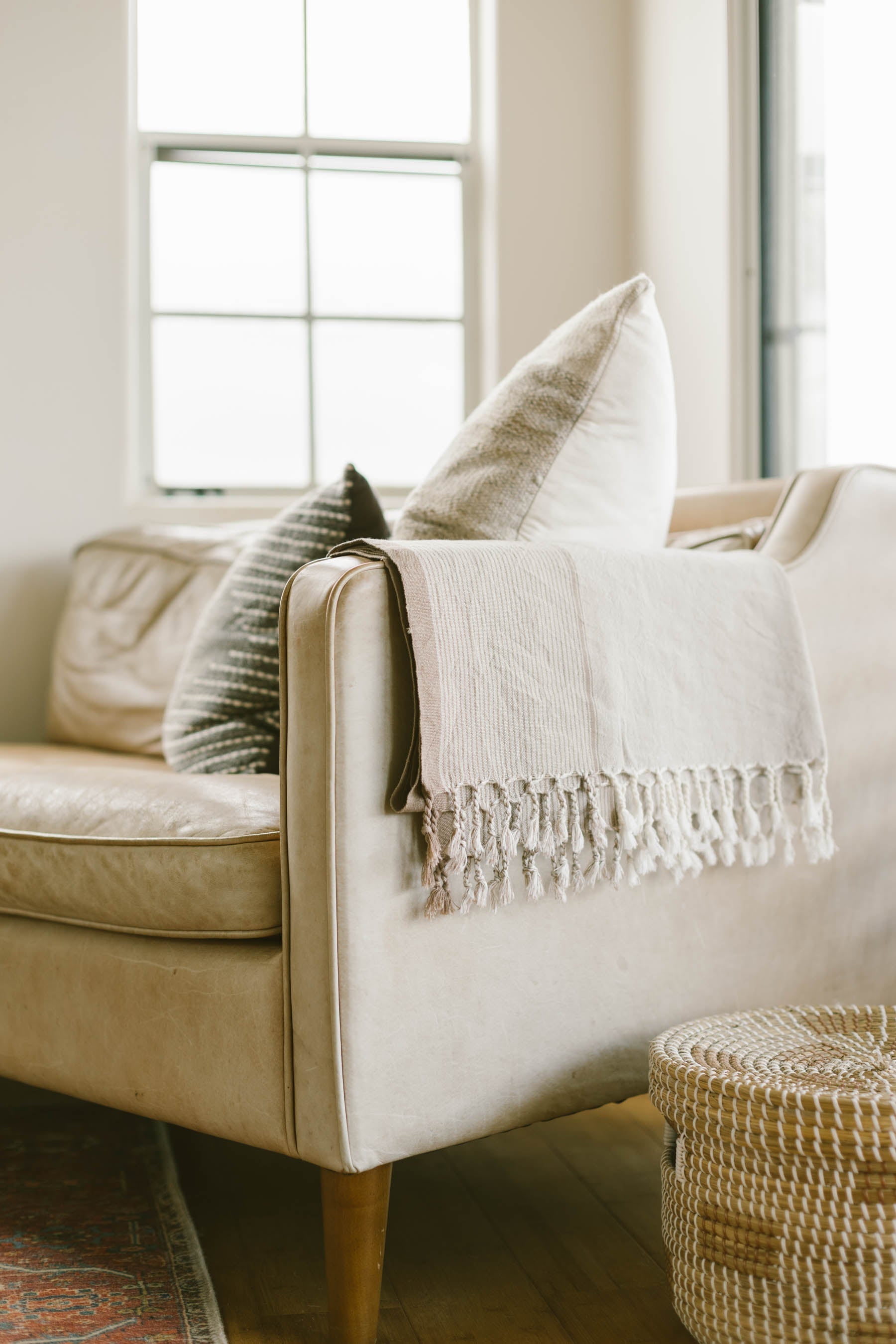 A beige Turkish towel with fringes is draped over the arm of a beige sofa, with a decorative pillow behind it and a woven basket on the floor beside the sofa