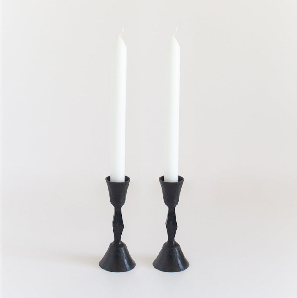 Two black, hand-forged iron candle holders with a textured finish, each holding a white candle.