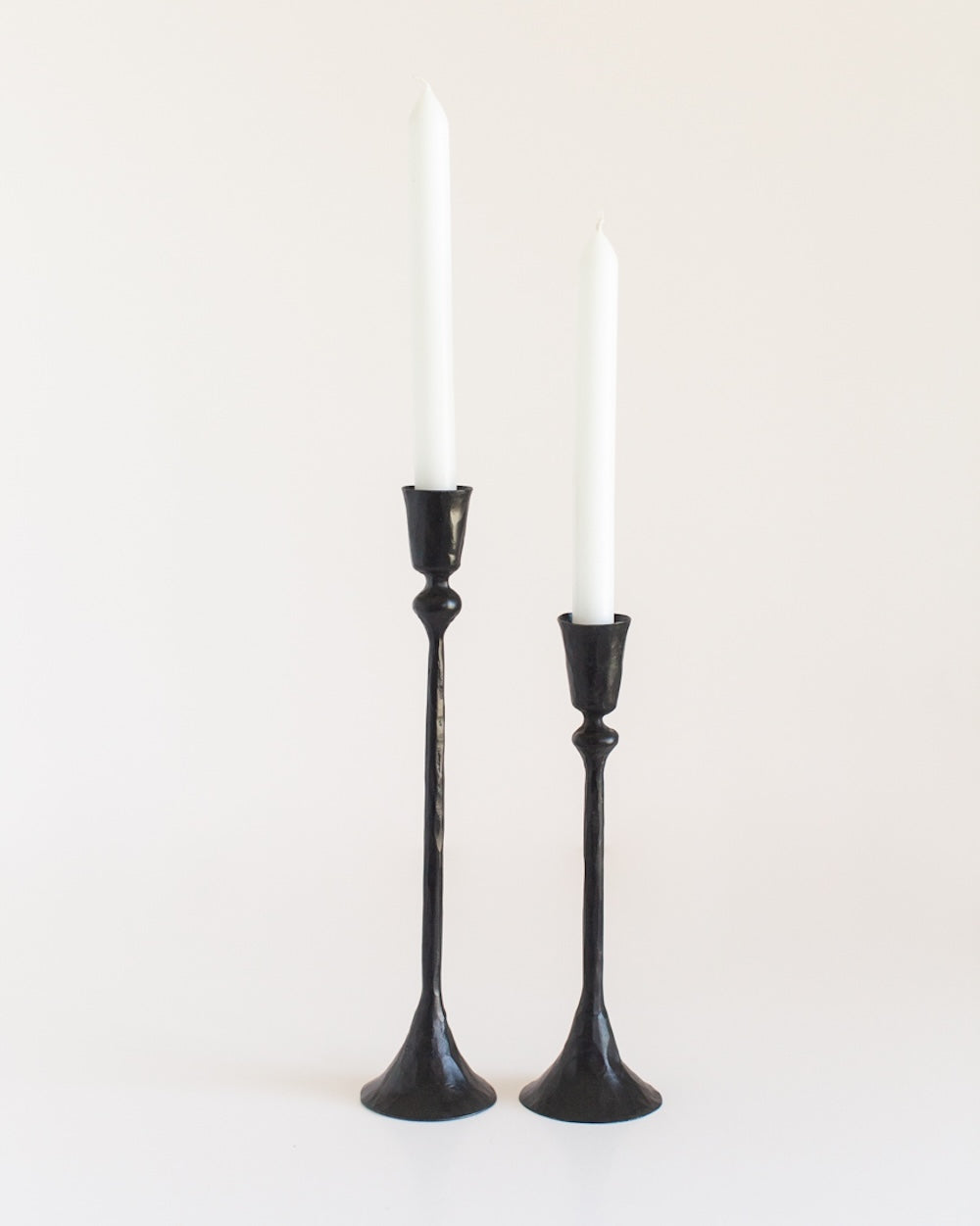 A pair of black, hand-forged iron candlesticks with a textured finish holding white candles, on a white background