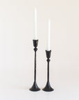 A pair of black, hand-forged iron candlesticks with a textured finish holding white candles, on a white background