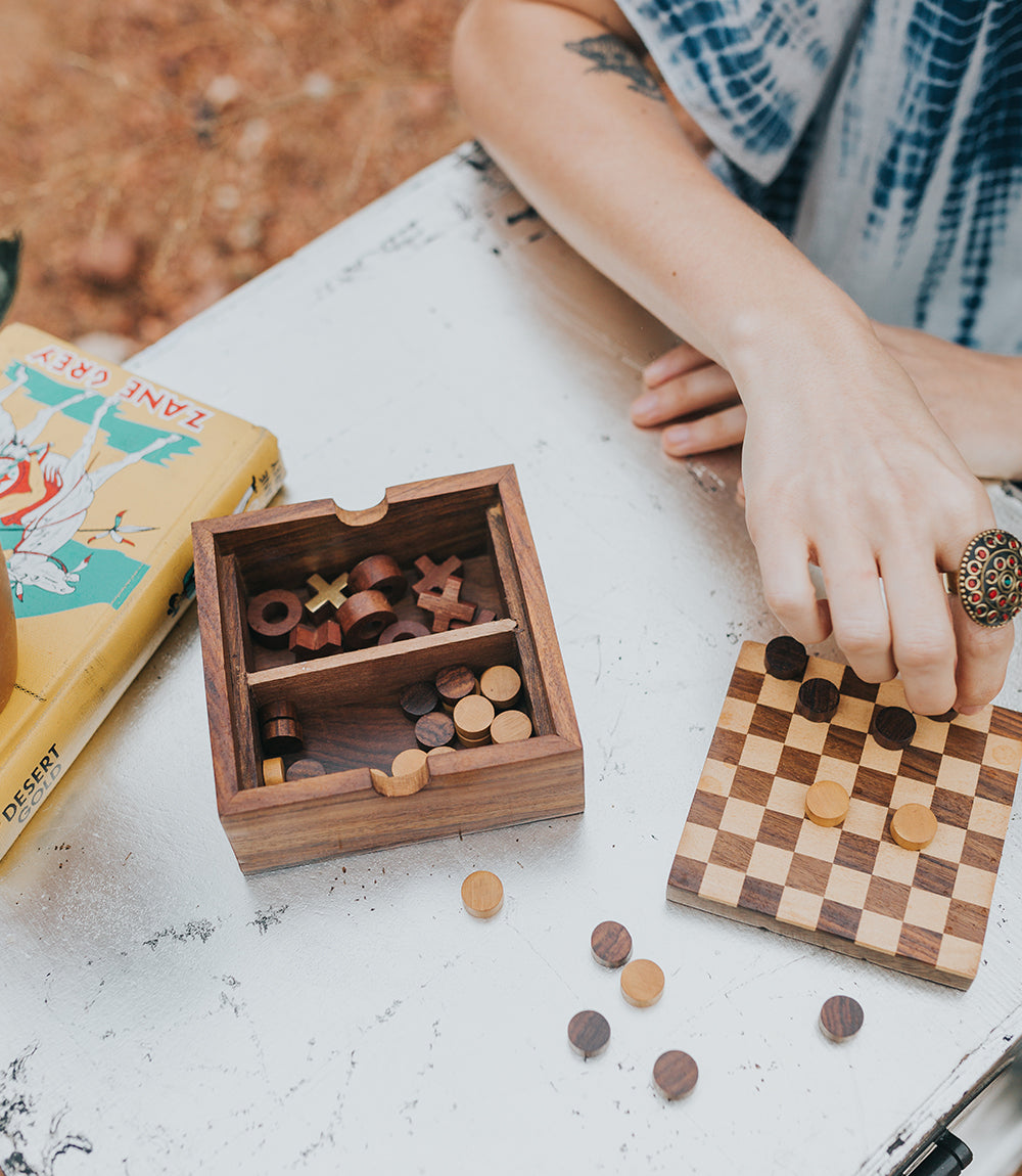A person playing a wooden 2-in-1 game set featuring Checkers and Tic Tac Toe on a reversible board with game pieces stored inside the compartments on a white table.