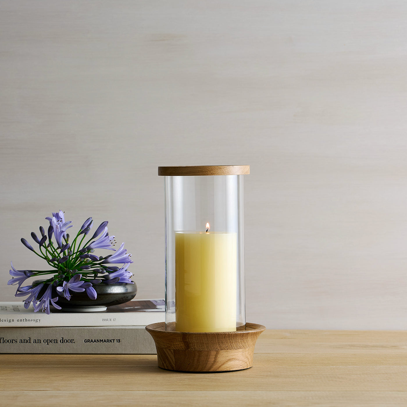 Candle in a glass holder with wooden base on a table with books and flowers.
