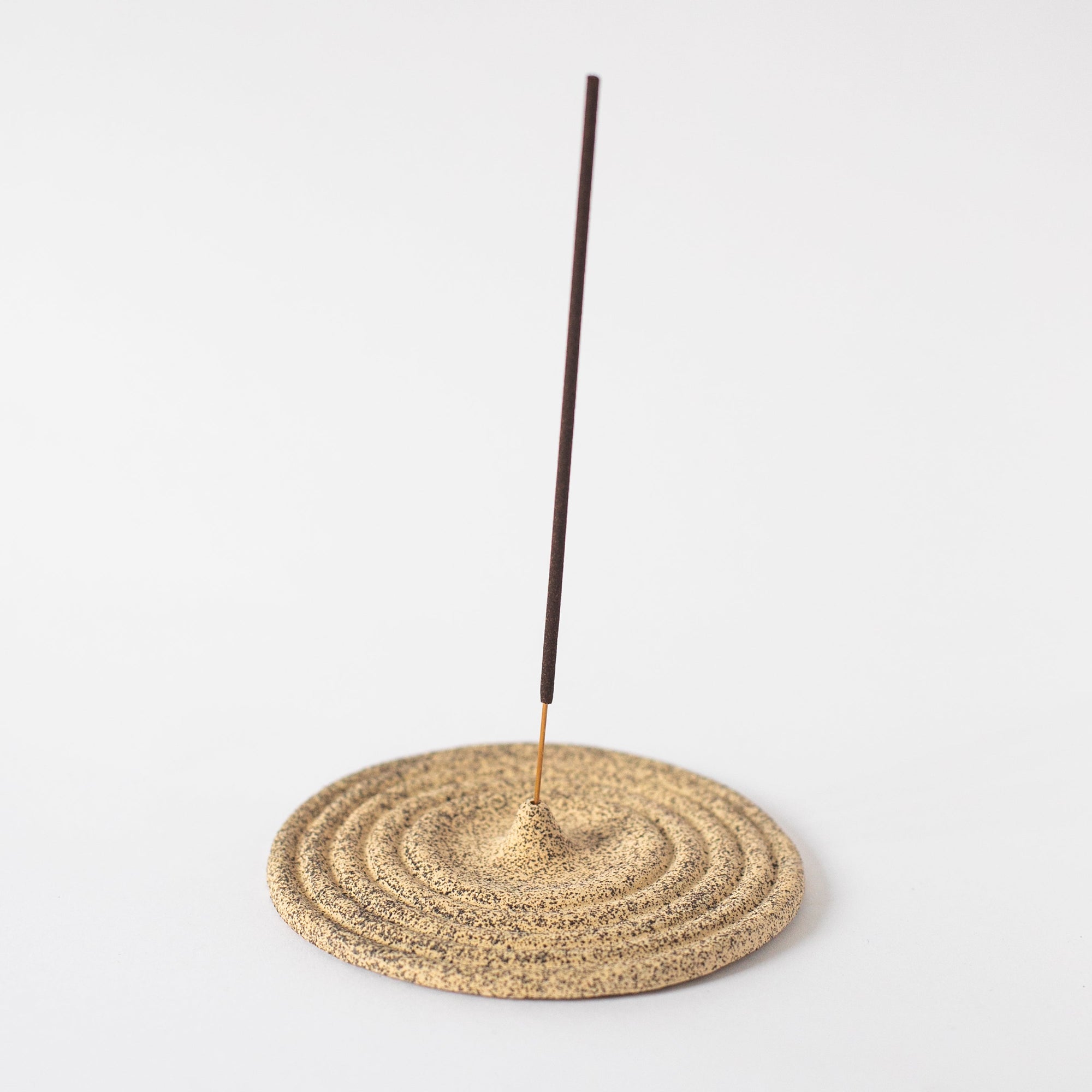 A beige ceramic incense holder with a rough texture, circular in shape, with an incense stick placed on top.