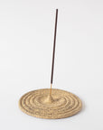 A beige ceramic incense holder with a rough texture, circular in shape, with an incense stick placed on top.