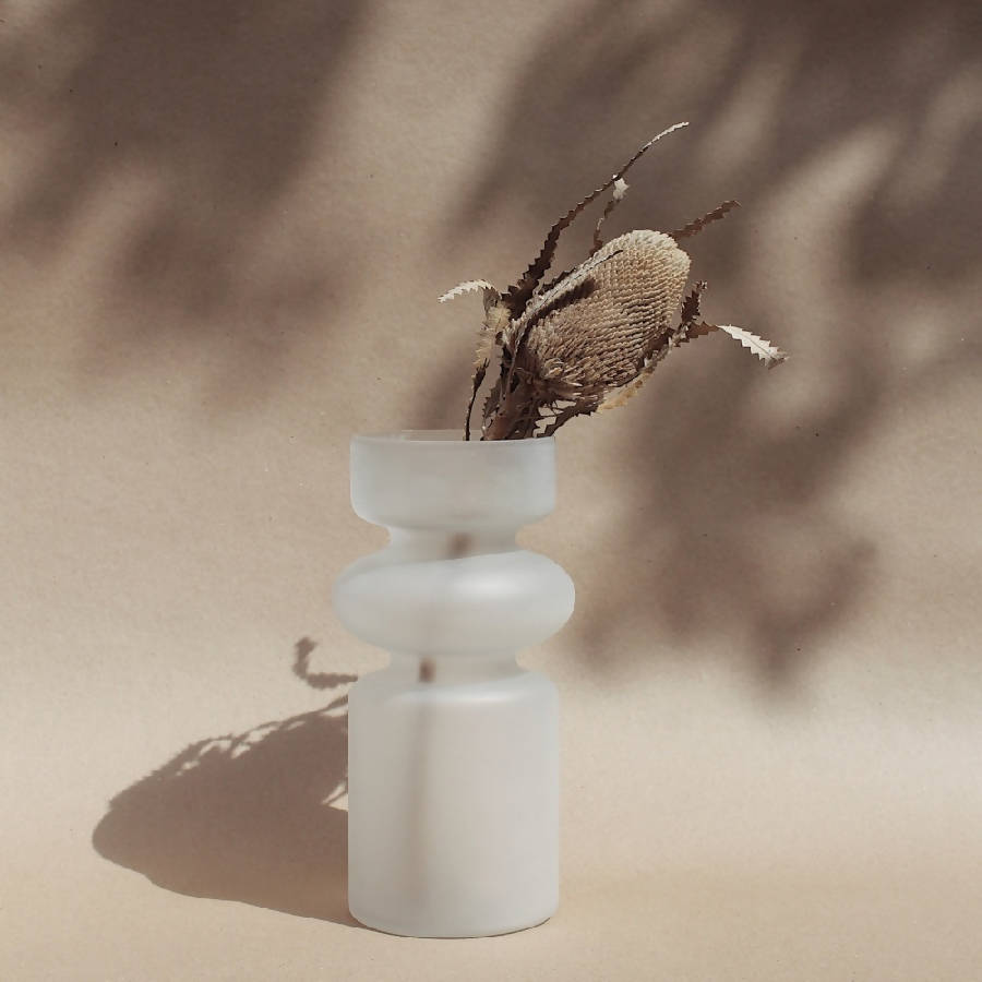 A frosted white glass vase with a translucent finish, featuring a symmetrical design, filled with dry twigs, positioned against a neutral background.