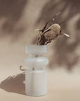 A frosted white glass vase with a translucent finish, featuring a symmetrical design, filled with dry twigs, positioned against a neutral background.