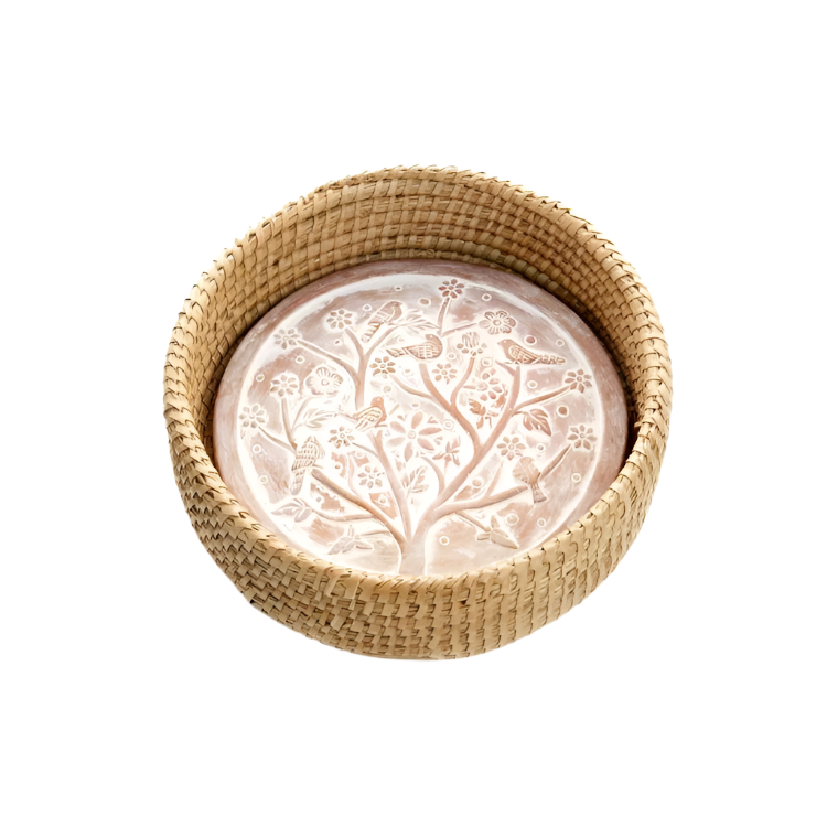 A whitewashed terracotta bread warmer with a birds of a feather design, placed inside a handwoven kaisa grass basket with palm wrap detail.