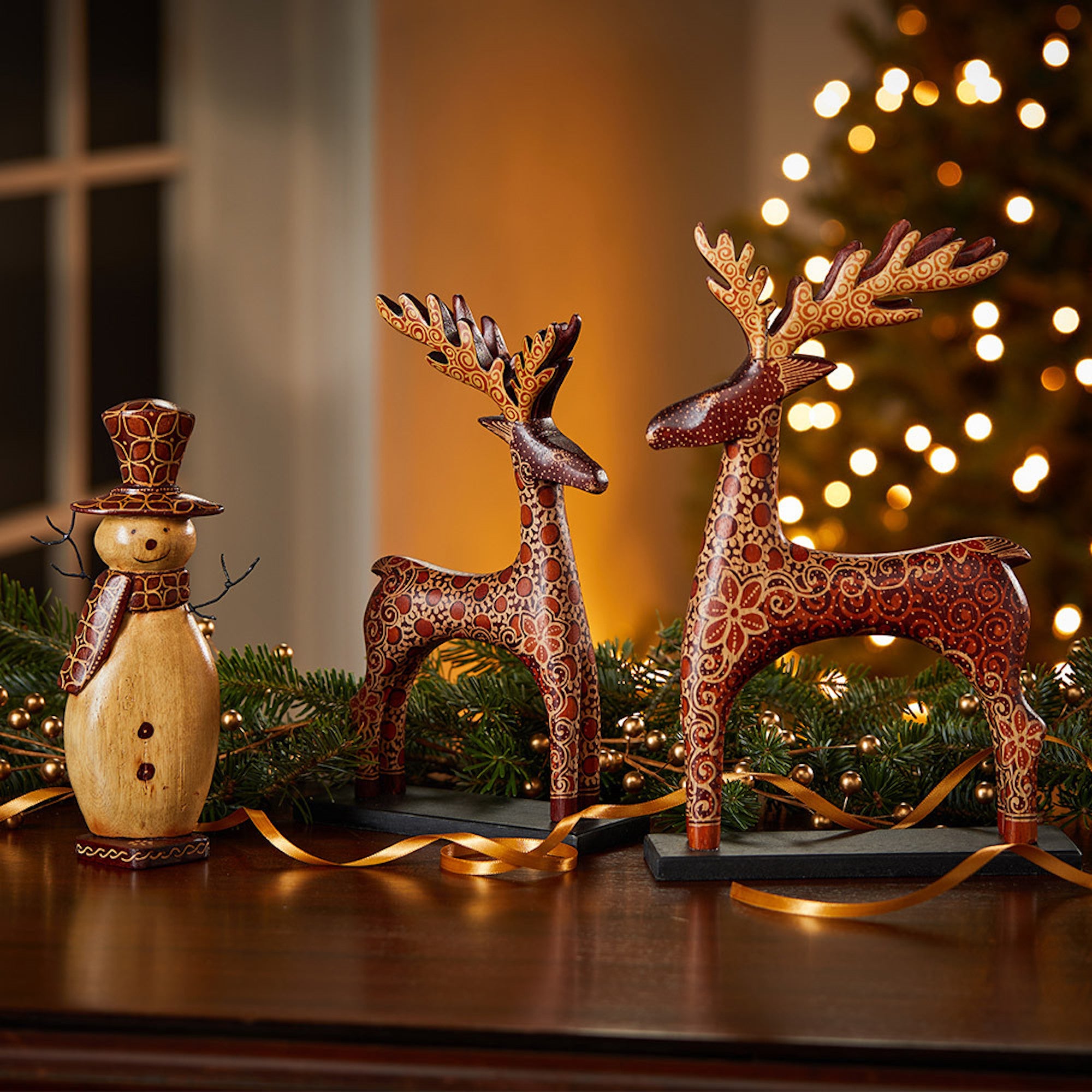 Decorative reindeer and snowman figures on a wooden surface with a blurred Christmas tree in the background.