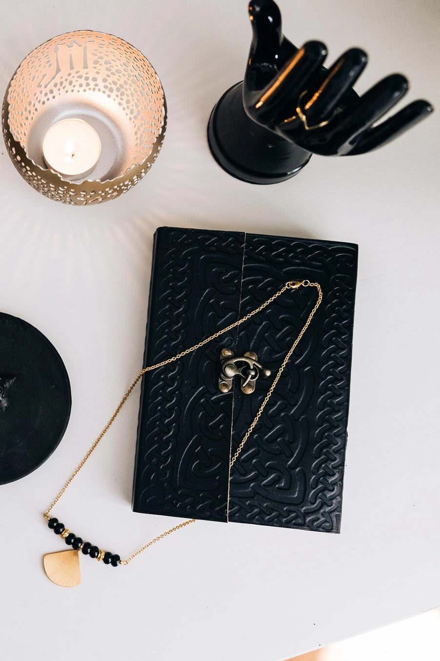 Black textured notebook with a gold necklace on a white surface with decorative items.