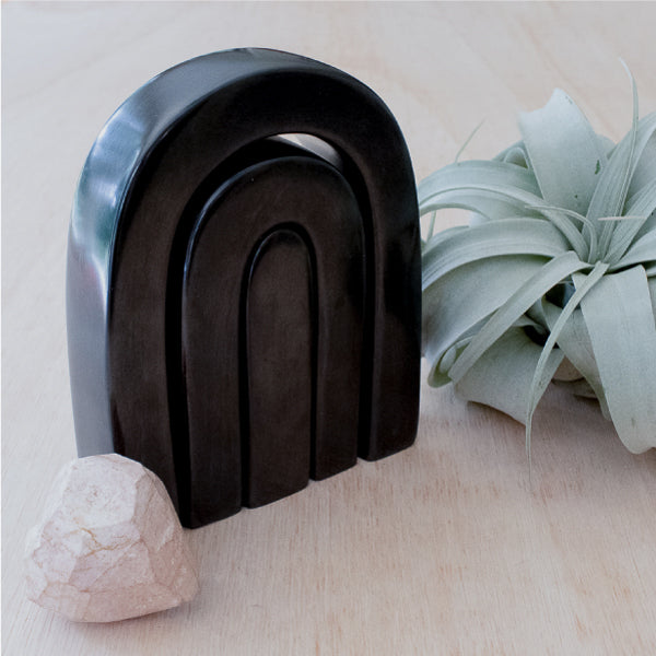 Set of three black arch-shaped objects on a light surface with a plant and rock in the background