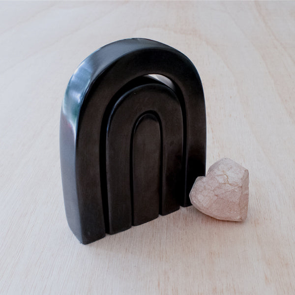 A stone black rainbow sculpture on a light wooden surface next to a heart shaped stone