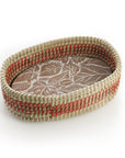 An oval terracotta bread warmer with a falling leaves design, featuring a handwoven kaisa grass basket with red dyed palm wrap detail.