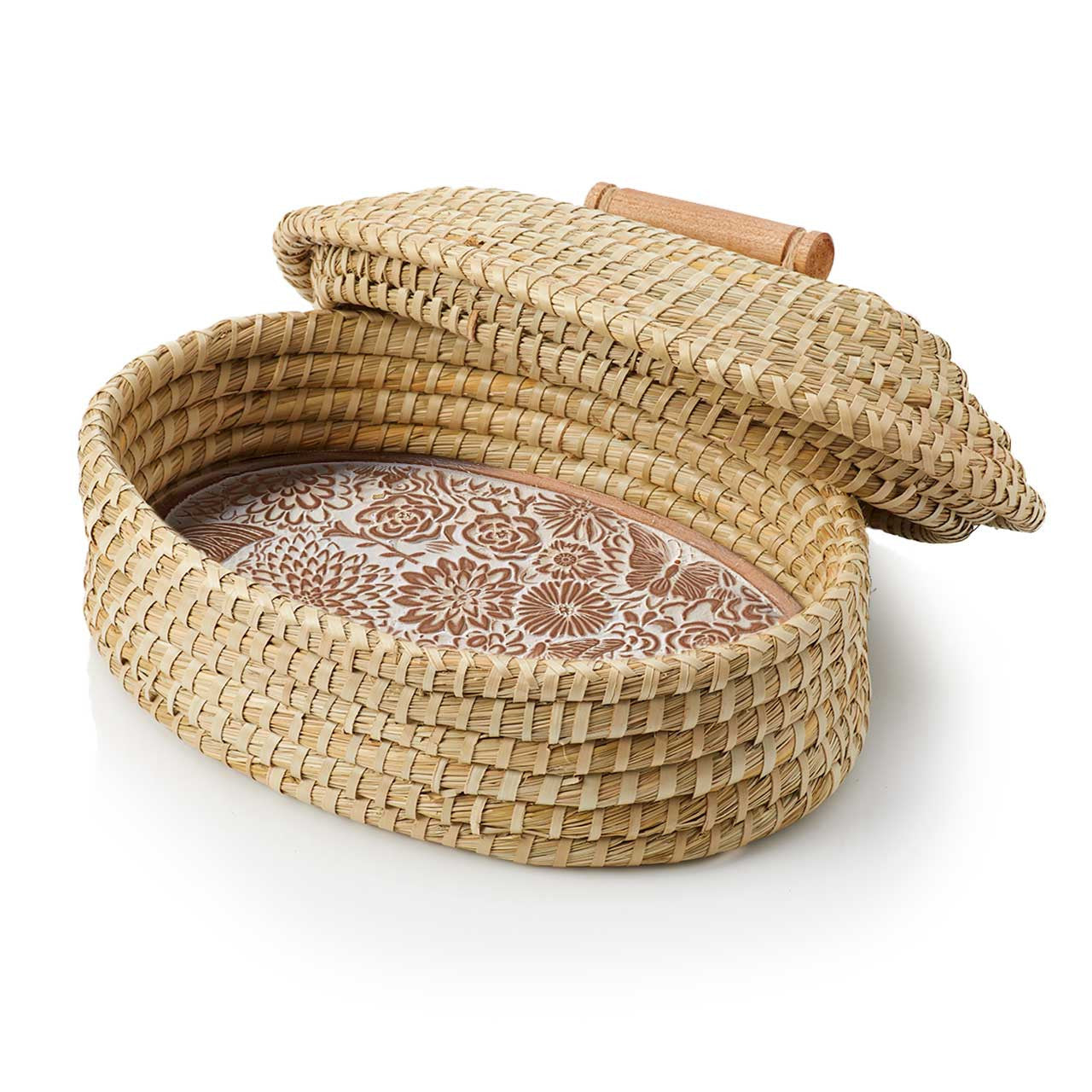 A terracotta bread warmer with a butterfly garden design, consisting of a stone incised with a pattern, placed inside a woven grass basket with a palm wrap detail, accompanied by a lid.