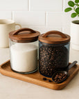 Glass jars with wooden lids containing sugar and coffee beans on a wooden tray.