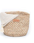 A beige handwoven bread basket with a napking hanging inside and a label showing the brand name 'Tallo de Olivo'