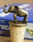 A brass elephan wine bottle stoppers sitting atop corks standing upright on top of a wooden table in front of a book.