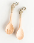 Two light natural wood serving utensils with elephant-shaped handles, one a salad spinner and one a spoon, displayed on a clean white background.
