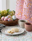 Easter eggs in a nest with floral patterns, two mugs, and cookies on a tablecloth.