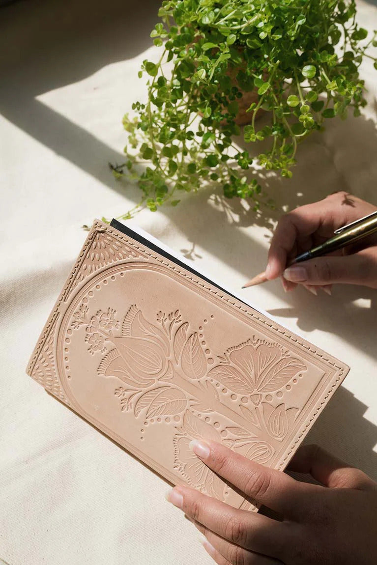 Person writing in a leather notebook with floral embossing on a light surface with a plant in the background