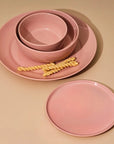 A set of pink dinnerware including one large plate, one medium plate, one large bowl, and one medium bowl, all with a wide rim