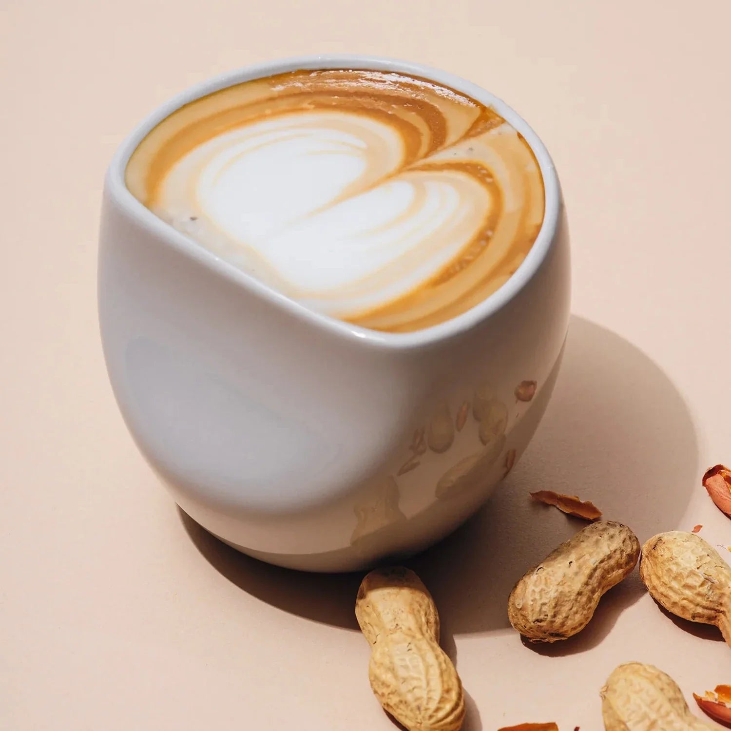 A large white ceramic coffee cup with a latte art design, accompanied by peanuts on the side, presented on a neutral background