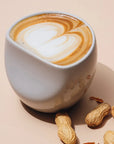 A large white ceramic coffee cup with a latte art design, accompanied by peanuts on the side, presented on a neutral background